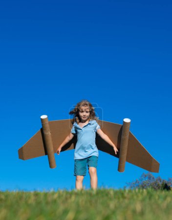 Foto de Pequeño niño juega astronauta o piloto. Niño en el fondo del cielo azul. Niños con alas de papel jetpack sueños. Los niños imaginan sueños de volar. Divertido niño con juguete jet pack - Imagen libre de derechos