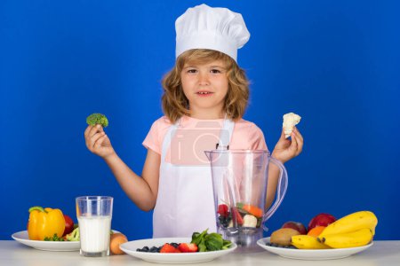 Téléchargez les photos : Garçon enfant dans le chapeau de chef et tablier tenir brocoli cuisson préparer le repas. Petit cuisinier avec des légumes à la cuisine. Alimentation naturelle pour enfants - en image libre de droit