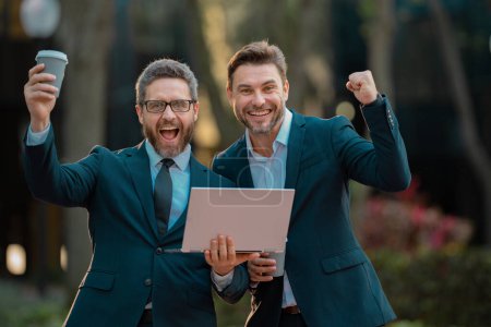 Photo for Two excited business men team using laptop outdoor. Amazed businessmen looking laptop with their business success in the city background. Two handsome young businessmen in classic suits using laptop - Royalty Free Image