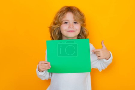 Téléchargez les photos : Enfant portant l'index sur une feuille de papier verte vide, isolé sur fond jaune. Portrait d'un enfant tenant une pancarte vierge, affiche - en image libre de droit
