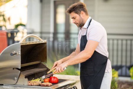Photo for Man cooking meat on barbecue in the backyard of the house. Handsome man preparing barbecue. Barbecue chef master. Man in apron preparing delicious grilled barbecue food, bbq meat. Grill and barbeque - Royalty Free Image