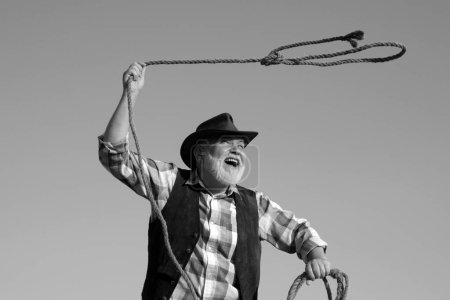 Foto de Viejo vaquero salvaje del oeste con cuerda. Hombre occidental barbudo lanzando lazo con chaqueta marrón y sombrero de captura de caballo o vaca - Imagen libre de derechos