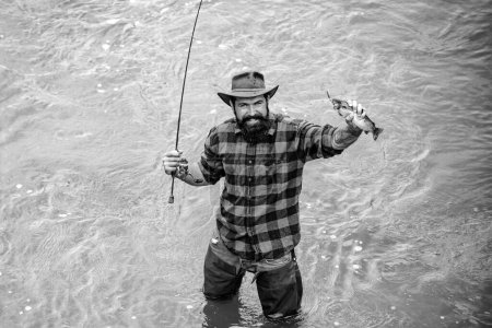 Foto de Fisher tiene pescado y caña larga. Hombre relajante y la pesca en el río. Feliz pescador barbudo en el agua. Pasándola bien. Día soleado activo. Hobby y actividad deportiva. Pasatiempo masculino - Imagen libre de derechos