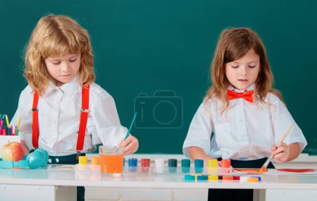 Téléchargez les photos : École enfants fille et garçon peinture avec des peintures couleur et pinceau dans la salle de classe. Enfant art, créativité enfant concept - en image libre de droit