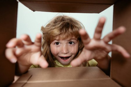 Téléchargez les photos : Drôle excité enfant déballer et ouvrir la boîte en carton regardant à l'intérieur. Le petit cadeau de déballage des enfants vue de l'intérieur. Enfant garçon regardant surpris dans un cadeau - en image libre de droit