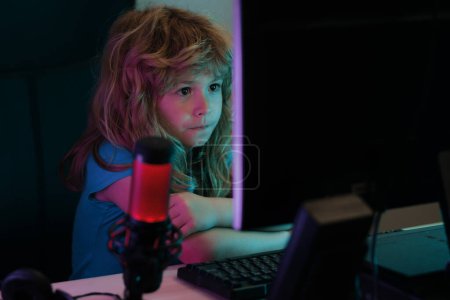 Téléchargez les photos : Petit gamer enfant jouer jeu d'ordinateur sur le bureau. Flux de jeu et blogging pour les enfants. École, étude, concept d'apprentissage en ligne - en image libre de droit