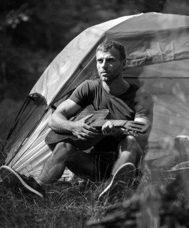 Photo for Young man sitting near touristic tent and playing guitar. Romantic time in a mountain camp. Tourism and hike, travel lifestyle. Handsome attractive model man playing guitar in forest with bonfire - Royalty Free Image