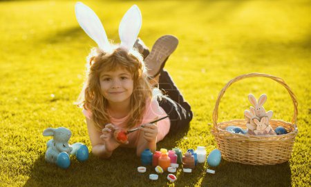 Foto de Niño con huevos de Pascua y orejas de conejo que ponen en la hierba pintando huevos. Feliz cara de niños de Pascua - Imagen libre de derechos