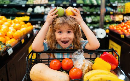 Photo for Child choosing a apple in a store. Funny little child smile and holding apple. Kid shopping in supermarket. Little boy with cart choosing fresh vegetables in local store - Royalty Free Image