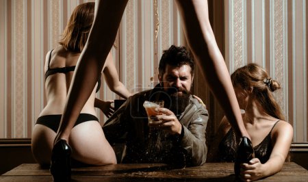 Téléchargez les photos : Sexy femme chaude avec de belles longues jambes debout à la table et de dessiner de la bière pour bel homme barbu à la maison publique. Homme barbu buvant de la bière et entouré de femmes chaudes sexy - en image libre de droit