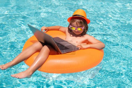Téléchargez les photos : Travailler et se détendre sur la plage. Enfant relaxant dans la piscine avec ordinateur portable. Enfant travaillant en ligne sur ordinateur portable, nageant dans une piscine d'eau turquoise ensoleillée - en image libre de droit