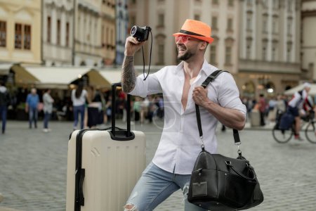 Foto de Hombre de turismo en jeans sombrero de moda y gafas de sol con maleta caminando por la calle. Foto de viaje del hombre con bolsa de viaje. Hombre con maleta sueña con viajar, aventura, vacaciones - Imagen libre de derechos