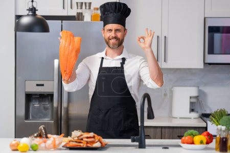 Photo for Portrait of man in chef apron and cook hat preparing fresh natural meal salmon at kitchen. Handsome cheerful chef man preparing raw fish salmon. Healthy food, cooking concept - Royalty Free Image