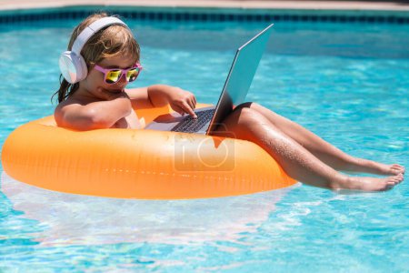 Téléchargez les photos : Enfant relaxant dans la piscine avec ordinateur portable. Enfant en ligne travaillant sur ordinateur portable, nageant dans une piscine d'eau turquoise ensoleillée. Voyage d'été et concept d'affaires - en image libre de droit