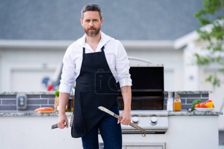 Téléchargez les photos : Un bel homme qui prépare un barbecue. Cuire la viande masculine sur le barbecue grill. Guy cuisinant de la viande sur le barbecue pour le dîner familial d'été dans la cour arrière de la maison - en image libre de droit