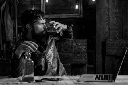 Foto de Hombre borracho. El hombre con barba sostiene brandy de vidrio. Un hombre sosteniendo un vaso de whisky. Bebiendo whisky. Degustación, degustación. Macho está bebiendo whisky durante el trabajo en su portátil - Imagen libre de derechos