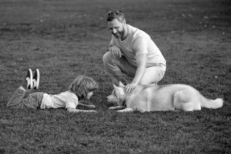 Téléchargez les photos : Père et fils sont assis sur l'herbe verte au parc d'été ou au jardin. Vacances en famille. Les animaux de compagnie aiment l'amitié et le concept des gens. Activité de week-end famille heureuse avec chien. Enfant avec ami animal - en image libre de droit