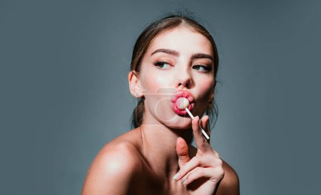 Photo for Girl licking candy. Lollipop model. Woman lips sucking a candy. Woman sucking lollipop in lips on grey background, isolated. Glamor beautiful young woman model with red lips eat sweats lolly pop - Royalty Free Image