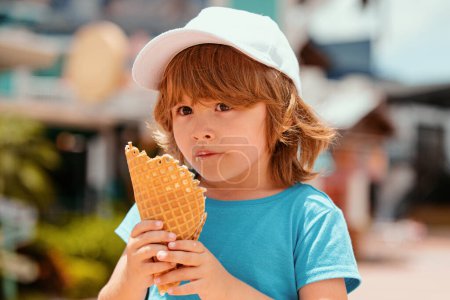 Téléchargez les photos : Un gosse qui mange de la glace. Joyeux petit garçon enfant manger de la crème glacée à l'arrière-plan de la ville - en image libre de droit