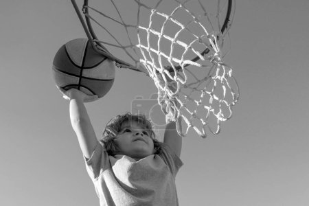 Téléchargez les photos : Basketball Slam Dunks de joueur de basket-ball pour enfants sportif. Image rapprochée du joueur de basket-ball enfant excité plongeant la balle, photo de stock. Bannière isolée sur fond de ciel - en image libre de droit