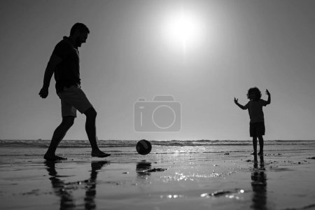 Foto de Padre e hijo juegan fútbol o fútbol en la playa en las vacaciones familiares de verano. Papá y el niño jugando al aire libre, silueta en la puesta del sol - Imagen libre de derechos