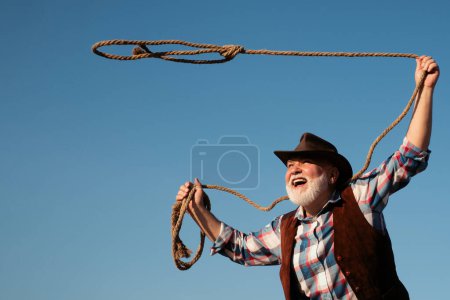 Foto de Viejo vaquero con cuerda de lazo en rancho o rodeo. Hombre occidental barbudo con chaqueta marrón y sombrero de captura de caballo o vaca - Imagen libre de derechos