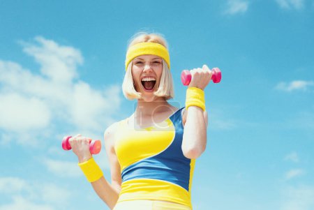 Foto de Atractiva joven fitness mujer sosteniendo dumbell. Mujer de fitness. Mujer en forma en la naturaleza en primavera. Feliz rubia levantando pesas - Imagen libre de derechos