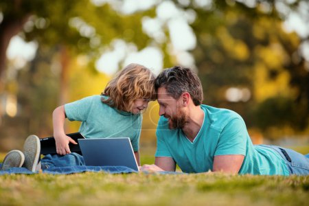 Photo for Weekend activity happy family lifestyle concept. Father with son use laptop. Dad and school boy child looking screen and tablet, watching video lesson, sitting on grass - Royalty Free Image