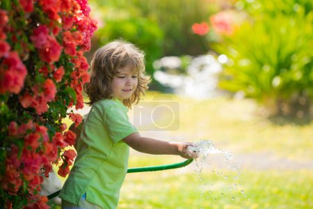 Foto de Niño jugando en el jardín, vierte de la manguera, hace una lluvia. Concepto de infancia feliz. Un niño derramando agua sobre los árboles. Cuidado de los niños para las plantas en el patio trasero - Imagen libre de derechos