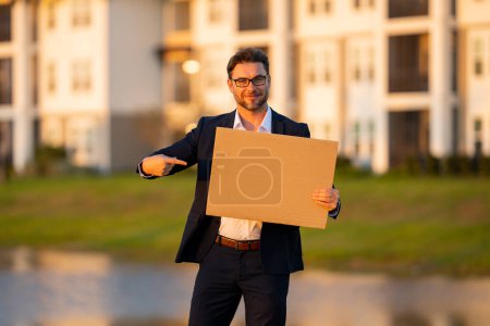 Photo for Business man with blank banner ad, posing outdoor. Portrait of attractive man in business suit holding empty blank poster. Man showing blank poster, pointing finger and gesturing index finger - Royalty Free Image