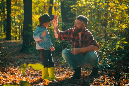 Foto de Padre e hijo jugando en el bosque de otoño. Niño pequeño con su padre hablando en otoño al aire libre. Elegante hombre con hijo, papá con joven hijo rubio en el parque de otoño - Imagen libre de derechos