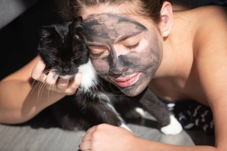 Téléchargez les photos : Bonjour avec le chat noir. Femme avec masque de boue faciale au fusain sur le visage. Procédure cosmétique. Spa de beauté et cosmétologie. Spa femme appliquant gris masque d'argile faciale. Soins de beauté - en image libre de droit