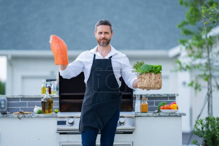 Téléchargez les photos : Un homme au tablier de chef tient des poissons de saumon au barbecue. Cuisinier masculin préparant barbecue à l'extérieur. Bbq poisson, grill pour pique-nique. Du saumon rôti. Homme attrayant préparant barbecue dans la cour de la maison - en image libre de droit