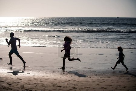 Téléchargez les photos : Joyeux jeune famille s'amuser sur la plage courir et sauter au coucher du soleil. Parents actifs et personnes en activité de plein air pendant les vacances tropicales d'été avec enfants - en image libre de droit