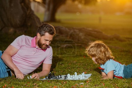 Téléchargez les photos : Père et fils jouant aux échecs allongés sur l'herbe à Lawn Park. Jeune garçon battant un homme aux échecs. Fête des Pères, famille amoureuse, parentalité, concept d'enfance - en image libre de droit