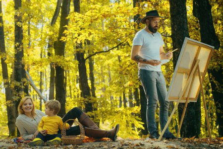 Foto de Hombre barbudo guapo dibuja un cuadro de su joven familia en el parque usando una paleta con pinturas y una espátula. Dibujo caballete y lienzo con un cuadro de personas estrechamente relacionadas - Imagen libre de derechos