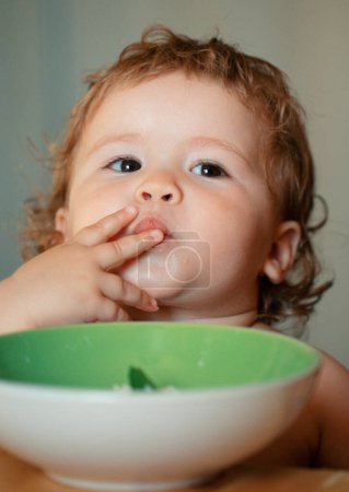Foto de Retrato de un niñito divertido comiendo de un plato sosteniendo la cuchara de cerca. Concepto de nutrición infantil - Imagen libre de derechos