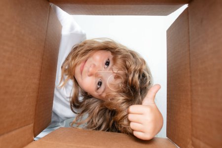 Foto de Divertido niño emocionado con el pulgar hacia arriba desempacar y abrir la caja de cartón mirando dentro. Los niños pequeños unboxing regalo dentro de la vista - Imagen libre de derechos