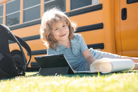 Téléchargez les photos : Elève écrit devoirs sur herbe avec tablette extérieure près du bus scolaire - en image libre de droit