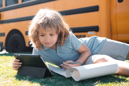 Téléchargez les photos : Étudiant écrire des devoirs sur l'herbe avec ordinateur portable en plein air près du bus scolaire. Concept d'enseignement à distance en ligne - en image libre de droit