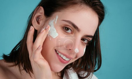 Téléchargez les photos : Belle jeune femme avec une peau propre et fraîche appliquant de la crème hydratante sur son joli visage - en image libre de droit