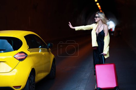 Téléchargez les photos : Une femme avec une valise prend un taxi. Taxi jaune. Voyageur femme touristique avec valise extérieure. Fille touristique avec voyage sac de voyage. Voyager en Europe le week-end - en image libre de droit