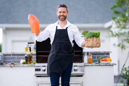 Téléchargez les photos : Homme en tablier de cuisine grillades et barbecue poisson de saumon dans le jardin. Un bel homme qui prépare un filet de saumon barbecue. Barbecue et grill. Cuisson des poissons dans la cour arrière - en image libre de droit