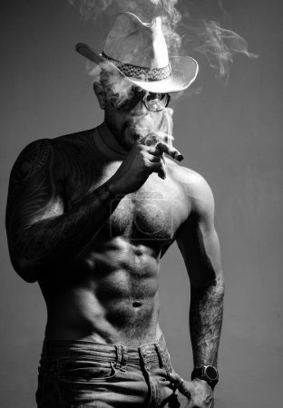 Téléchargez les photos : Santé. fumer une cigarette. expirez la fumée. mauvaise habitude. le charisme de confiance. abdos sexy de tatoueur. mode masculine. homme macho musclé avec corps athlétique. sportif brutal en chapeau de cow-boy - en image libre de droit