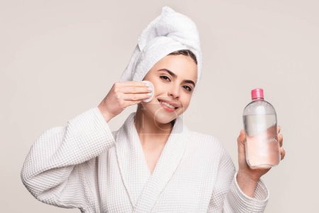 Téléchargez les photos : Joyeux jeune femme en peignoir avec serviette sur le visage de nettoyage de la tête avec de l'eau micellaire et coton isolée sur rose. Fille avec de l'eau micellaire - en image libre de droit