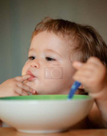 Photo for Funny little baby in the kitchen eating with fingers from plate. Lick tasty fingers - Royalty Free Image