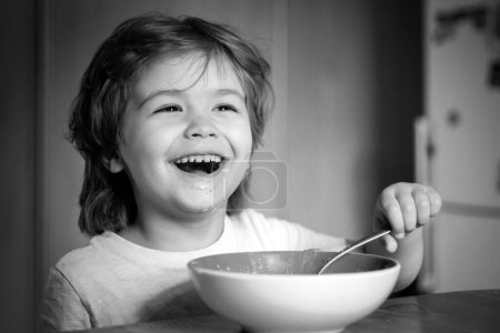 Téléchargez les photos : Un gosse qui mange. Petit garçon prenant son petit déjeuner dans la cuisine. Souriant heureux adorable bébé manger de la purée de fruits dans la cuisine. Petit garçon affamé mangeant - en image libre de droit