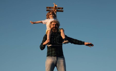 Photo for Father and son. Fathers day. Father giving son ride on back. Portrait of father giving son piggyback ride on shoulders and looking up. Cute boy with dad playing with wooden airplane outdoor - Royalty Free Image