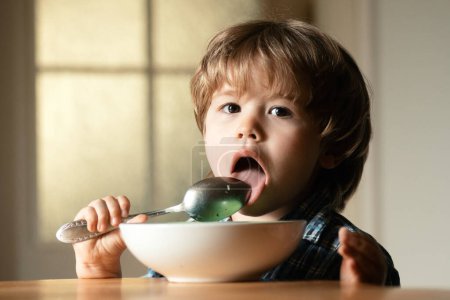 Photo for Young kid sitting on the table eating with funny expression on face. Tasty kids breakfast. Tasty kids breakfast. Cheerful baby child eats food itself with spoon - Royalty Free Image