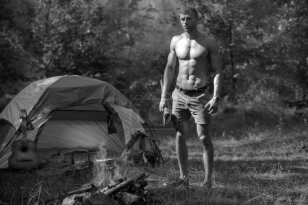 Foto de Temporada de camping con fogata. Hombre con torso desnudo, campamento de turismo de picnic. Hombre leñador con hacha mirar llama hoguera en el día de verano en el paisaje natural. Viajar, acampar vacaciones senderismo - Imagen libre de derechos